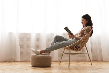 Relaxed girl reading book sitting in chair by the window