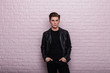 Fashionable attractive young man in black clothes in a stylish leather jacket is resting near a vintage pink brick wall in the room. Handsome modern trendy hipster guy is standing in the studio.