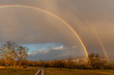 Fototapeta Tęcza - Feldweg mit doppeltem Regenbogen vor dunkler Wolkenwand 