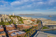 San Francisco, California, daytime, sunrise, aerial view of North Beach with Coit Tower and Golden Gate Bridge visible. Blue sky with clouds and golden light. Embarcadero area in foreground. 