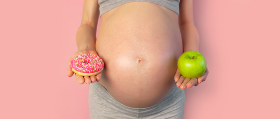 Beautiful young pregnant woman holding green apple and sweet donut in her hand on pink background. Expectation of the child, pregnancy and motherhood. Healthy and unhealthy eating concept, diet