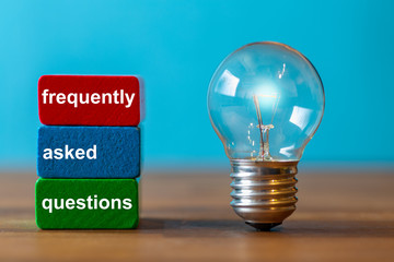 frequently asked questions are the word written on a red and a green toy block. Next to the tow blocks, an ancient light bulb with glowing light stands freely and upright on a dark wooden table.