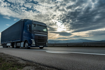 Blue semi trailer truck driving on a highway with dramatic sunset sky in the background. Transportation vehicle