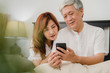 © tirachard - Asian senior couple using mobile phone at home. Asian Senior Chinese grandparents, husband and wife happy after wake up, watching movie lying on bed in bedroom at home in the morning concept.