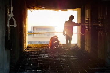 2 construction workers sitting and standing, wear helmet and working about steel ties to wait for the concrete on the building under construction. Window view, high rise condominium background, sunset