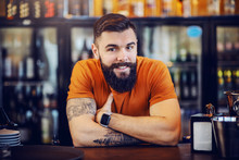 Portrait Of Handsome Bearded Smiling Positive Tattooed Barman Leaning On Bar Counter, Looking At Camera And Waiting For Customers To Order Drinks.