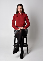 Poster - full length portrait of a pretty brunette girl wearing a red shirt and plaid skirt with leggings and boots. Sitting on a chair against a  studio background.