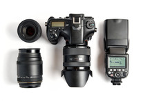 Top View Of Modern Digital Camera Equipment - DSLR With Attached Zoom Lens And Hood, Lenses And External Flashlight On White Background