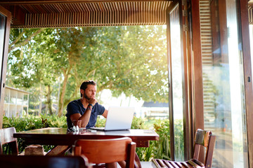 Freelance concept. Thoughtful Young  man using laptop while sitting on cafe terrace.