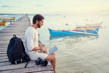 Work And Travel. Young Man With Rucksack Using Laptop Computer Sitting On Wooden Fishing Pier With Beautiful Tropical Sea View.