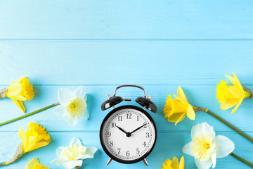 Black alarm clock with spring flowers and space for text on light blue wooden background, flat lay. Time change