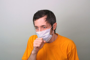 Wall Mural - Middle-aged adult man in a yellow T-shirt on a gray background in a medical mask sneezes covering his face with his hand. Cough, fever. Disease protection coronavirus epidemic problem from China