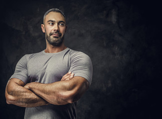 Strong, adult, fit muscular caucasian man posing for a photoshoot in a dark studio under the spotlight wearing grey sportswear, showing his muscles with arms crossed, looking confident and calm