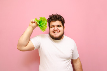 Wall Mural - Funny young overweight man isolated on pink background with green leaves in his hands, looks into the camera and smiles.Fat man on a diet, holding lettuce leaves, eating healthy food