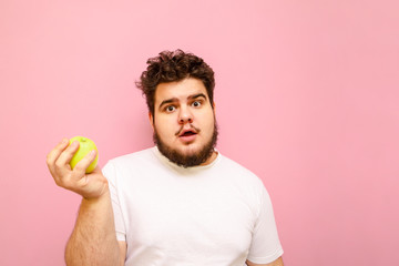 Wall Mural - Surprised young overweight man stands on pink background with green apple in hand, looks into camera with shocked face. Funny fat guy with a surprised face holds an apple in his hand. Isolated