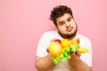 Wall Mural - Closeup portrait of a funny young man with beard and curly hair holding fruits and vegetables in his hand and looking into the camera. Fat young guy holding healthy food in his hand, diet concept.