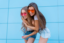 Two Young Hipster Woman Friends In Retro Neon Sunglasses Standing And Smiling Over Blue Wall