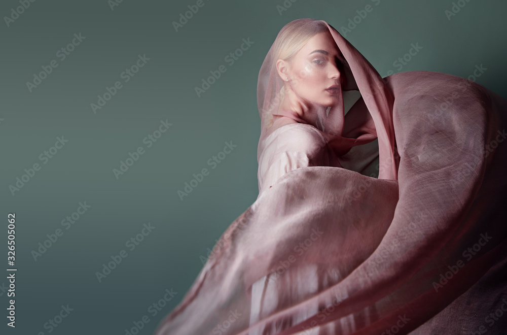 A beautiful young tender girl in a chiffon dress is covered with a drapery made of thin fabric that develops in the wind. - obrazy, fototapety, plakaty 