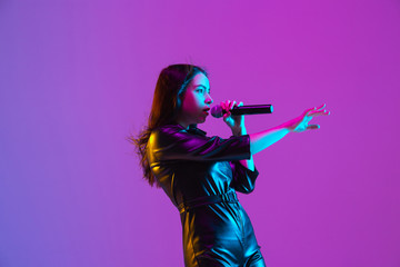 Wall Mural - Caucasian female singer portrait isolated on purple studio background in neon light. Beautiful female model in black wear with microphone. Concept of human emotions, facial expression, ad, music, art.