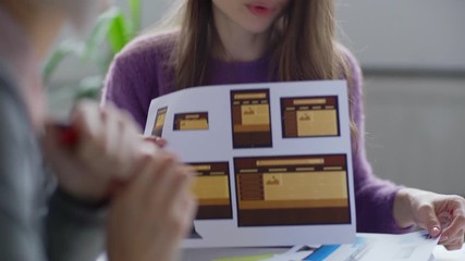 Wall Mural - Tilt up of young female designer showing print outs with website templates for different devices to colleague typing on laptop at office desk