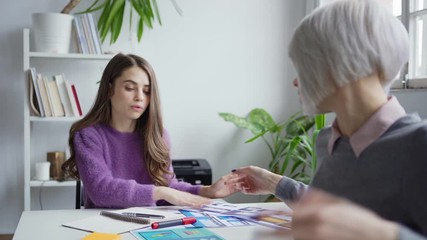 Wall Mural - Female designer sitting at table with open laptop, turning to busy colleague, clarifying details of project and discussing webiste interface templates printed on papers in office
