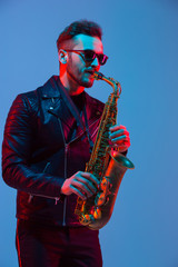 Wall Mural - Young caucasian jazz musician playing the saxophone on gradient blue-purple studio background in neon light. Concept of music, hobby, festival. Joyful attractive guy. Colorful portrait of artist.