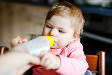 Wall Mural - Cute adorable baby girl holding nursing bottle and drinking formula milk. First food for babies. New born child, sitting in chair of domestic kitchen. Healthy babies and bottle-feeding concept