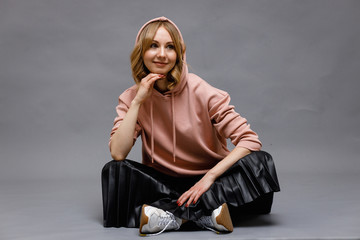 Wall Mural - A beautiful young girl sitting on the floor  and smiles in a pink hoodie, black skirt and white sneakers on a gray background.