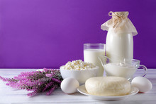 Glass And A Bottle Of Milk, A Plate Of Cottage Cheese, Cheese, White Raw Eggs, Sour Cream In A Gravy Boat, Lilac Lavender Flowers On A White Wooden Surface On A Purple