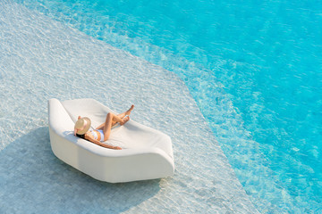 Top view of Asian woman wearing sexy white swimsuits with hat leisure and relaxing lounging on white outdoor sofa sun bed lounger at the big swimming pool. Summer vacation concept.