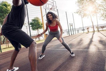 Outdoors Activity. African couple playing basketball on court dribbling girl looking camera playful