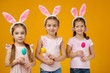 © producer - happy cute little child girls with pink bunny ears holding painted Easter eggs on studio yellow background. Easter day