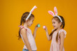 © producer - happy cute little child girls with pink bunny ears holding painted Easter eggs on studio yellow background. Easter day