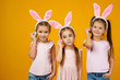 © producer - happy cute little child girls with pink bunny ears holding painted Easter eggs on studio yellow background. Easter day