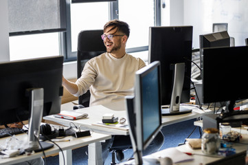 young programmer sitting at the desk in his office and working .he talk with someone and smiling.