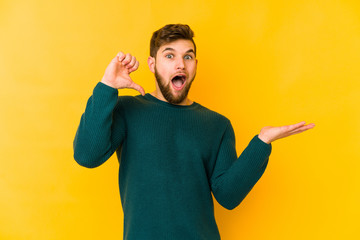 Wall Mural - Young caucasian man isolated on yellow background holds copy space on a palm, keep hand over cheek. Amazed and delighted.