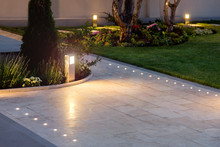 Marble Tile Playground In The Night Backyard Of Mansion With Flowerbeds And Lawn With Ground Lamp And Lighting In The Warm Light At Dusk In The Evening.