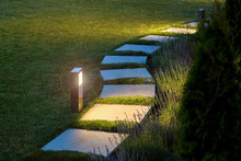 Marble Path Of Square Tiles Illuminated By A Lamp Glowing With A Warm Light In A Night Garden With A Flower Bed And A Lawn, Nobody.