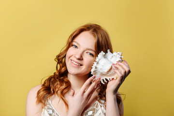 A young woman put a sea shell to her ear. A girl with red hair smiles and looks at the camera, listening to the sound of the sea in a sea shell, standing against the wall on a yellow background.