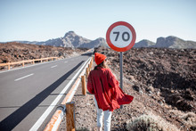 Lifestyle Portrait Of A Woman In Red Walking On The Roadside Near The Huge Road Sign, While Travel On The Volcanic Valley. Carefree Lifestyle And Travel Concept