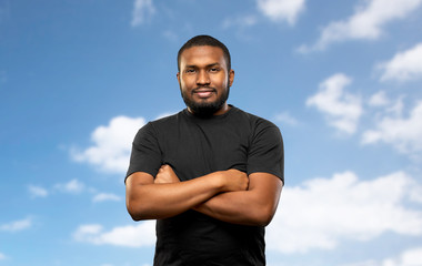 Wall Mural - people concept - young african american man in black t-shirt with crossed arms over blue sky and clouds background