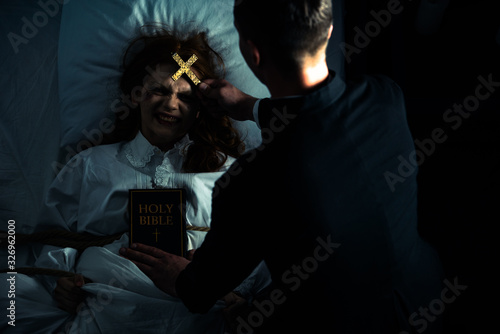 Fotografía exorcist with bible and cross standing over demonic obsessed girl in bed