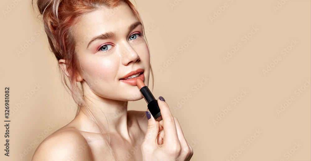 Portrait of gorgeous smiling woman holding rosy lipstick in hand. Model posing in studio and looking in camera gracefully. Beauty and cosmetics concept. Isolated on beige - obrazy, fototapety, plakaty 