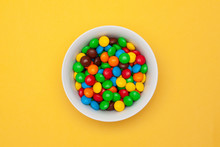 White Bowl With Chocolate Colored Round Candy On Yellow Background In The Center. Top View.
