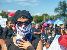 A Girl With Glasses, Hat And Scarf Covering Her Mouth, Fights For Her Rights With The Dominican Flags And The Blue Sky In The Background