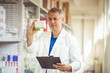 Smiling handsome male pharmacist promoting a product holding it in his hand visible to the camera with a friendly smile. Smiling male pharmacist in white coat over drugstore background