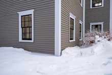 A Tan Colored Vintage Wooden Building With Multiple Windows. The Exterior Walls Are Covered In Narrow Clapboards And Wide White Corner Boards. There's A Drift Of Snow In Front Of The Building. 