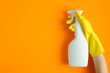 Hand in yellow rubber glove holding plastic spray bottle with cleaning detergent on a orange background