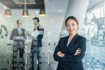 Wall Mural - Portrait of young asian businesswoman standing at her office looking at camera. background of businessman.