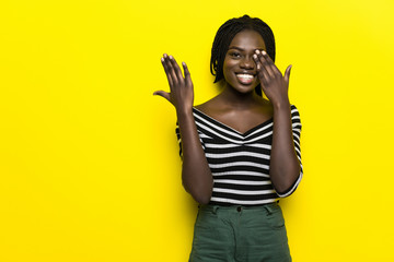 Wall Mural - Beauitul african american woman peeking in shock covering face and eyes with hand, looking through fingers with embarrassed expression over isolated yellow background